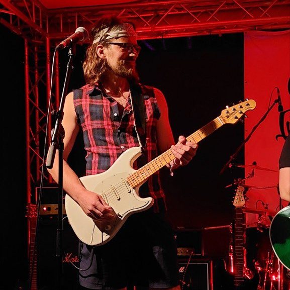 Musiker mit weißer Gitarre auf der Bühne, trägt ein kariertes Hemd ,Brille und Stirnband