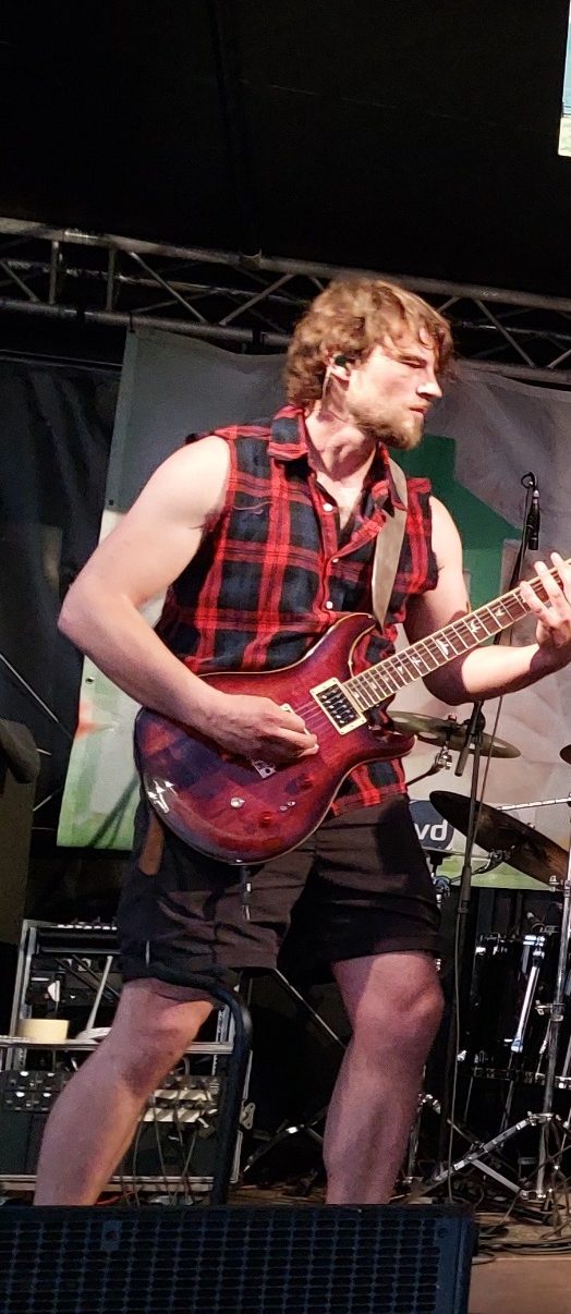 Mann mit blonder Kurzhaarfrisur, der auf einer Gitarre spielt, trägt ein kariertes Hemd.