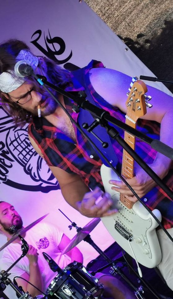 Mann spielt Gitarre auf der Bühne, mit Bandana und kariertem Hemd, während ein Schlagzeuger spielt.