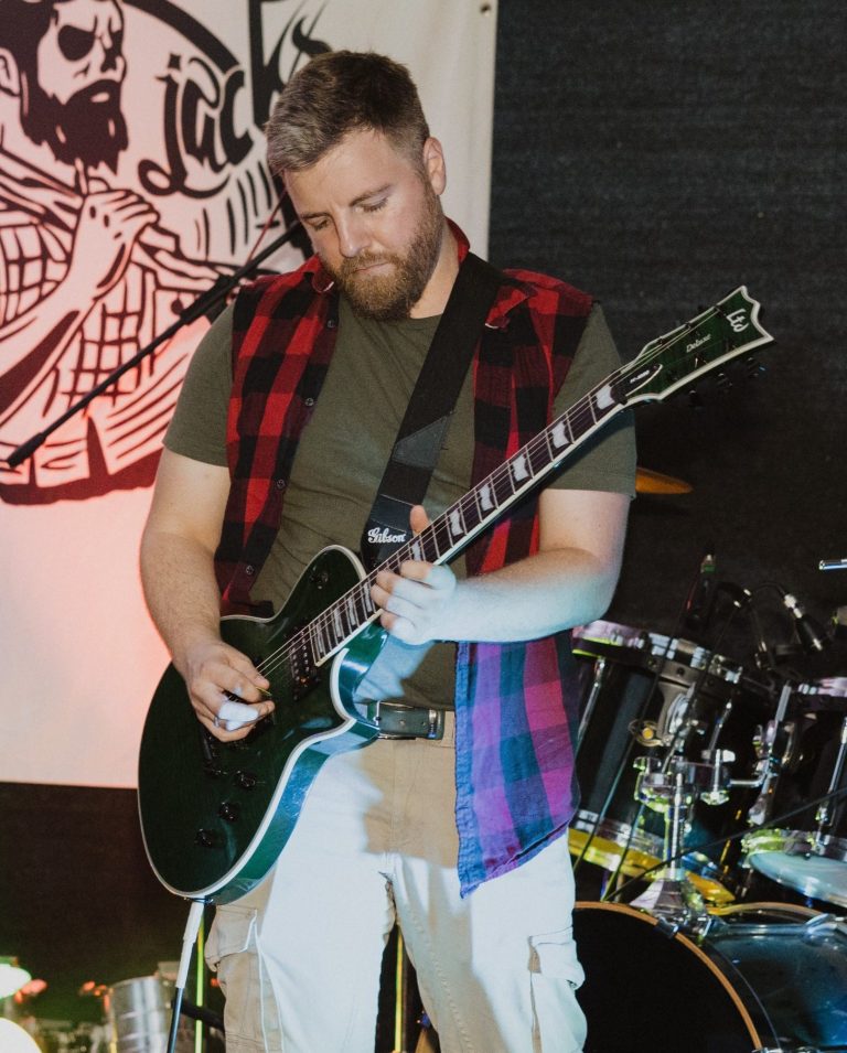 Mann spielt E-Gitarre auf einer Bühne, im Hintergrund Schlagzeug ,Logo der Band und Bühnenbeleuchtung.