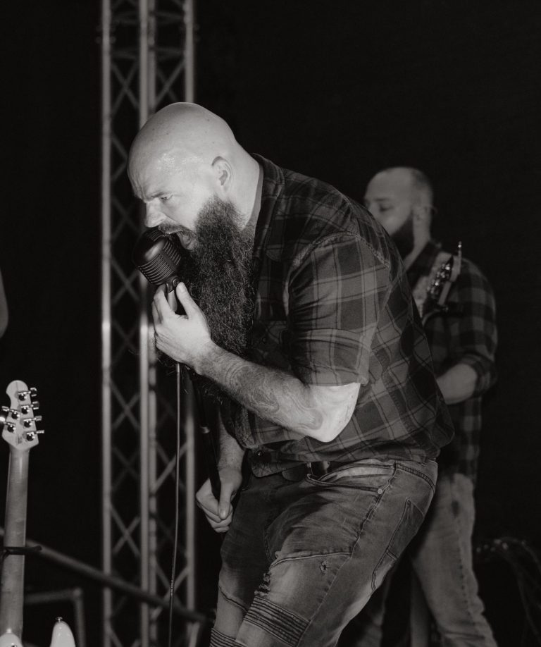 Mann mit Bart und Glatze singt in ein Mikrofon auf der Bühne. Bassist im Hintergrund. 