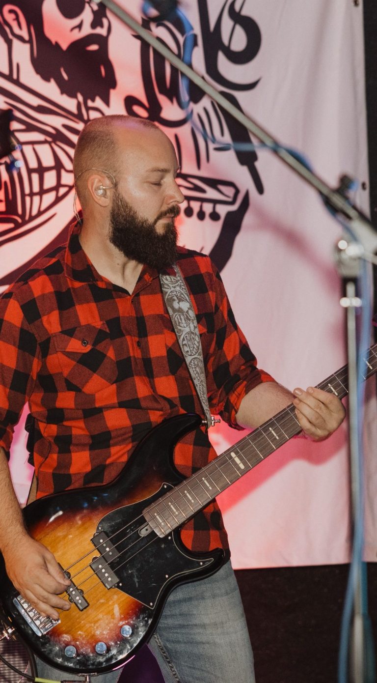 Mann mit Bart spielt E-Bass auf einer Bühne, im Hintergrund ein kreatives Bandlogo.